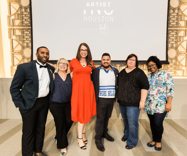 Group of artists dressed in business casual and formal attire pose together in front of a projection screen.