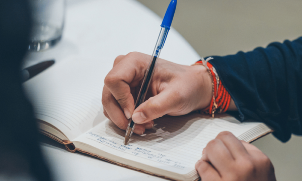 Person's arm and hand holding a pen and writing on a pad of paper.