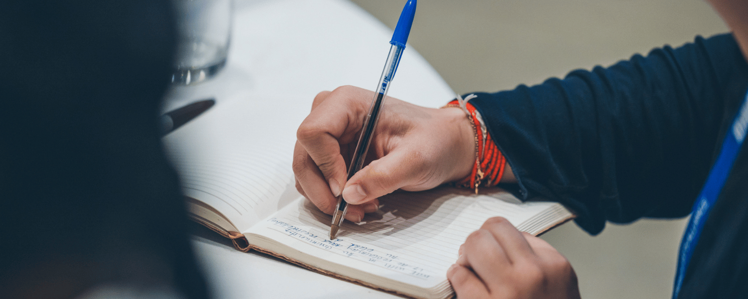 Person's arm and hand holding a pen and writing on a pad of paper.