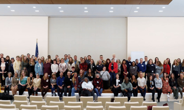 A large group of people pose for a photo in a bright auditorium with rows of white seats. Most are standing, while a few are sitting on tiered platforms. An American flag is visible in the background. The setting is clean and professional.