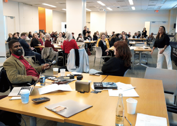 A diverse group of people seated at tables in a conference room, attentively facing a presenter at the front. Papers, bottles, and electronic devices are on the tables. The room is well-lit and spacious, with a collaborative atmosphere.