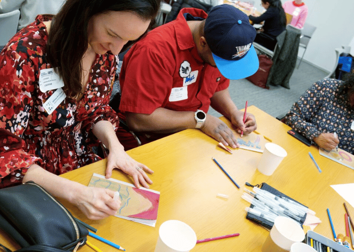 Three people are sitting around a table engaged in drawing with colored pencils. They are each focused on their individual artwork. Various art supplies are scattered on the table, including pencils and markers. Cups and bags are also visible.