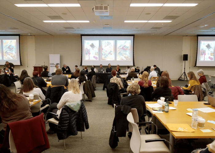 A conference room filled with people seated at tables facing a panel of speakers. Three screens display the same colorful graphic. Attendees are mostly women, and some have laptops open on the tables.