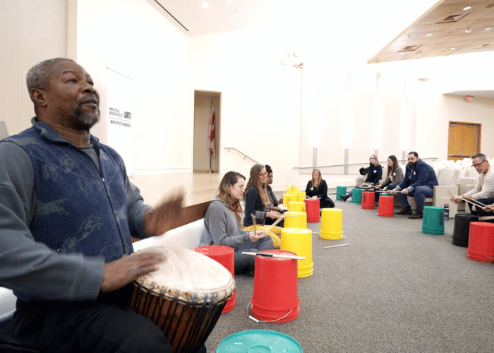 A group of people sit in a semi-circle, each playing colorful drum buckets with sticks. A man on the left plays a traditional drum. The room is bright with a minimalist design, and everyone appears engaged and focused.