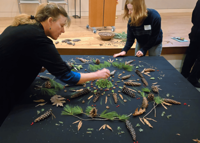 Two women lean over a table arranging leaves, pine branches, and pine cones into a mandala shape.