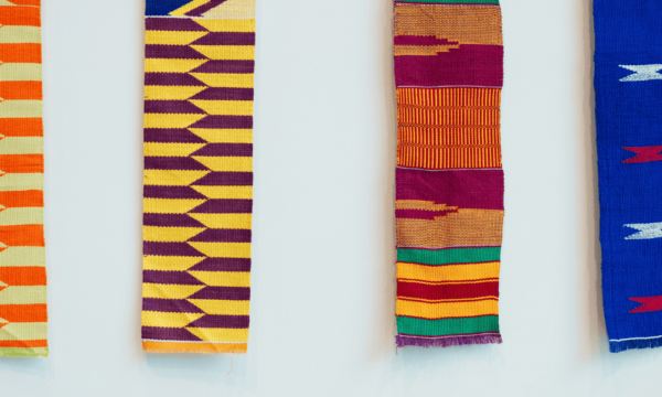 Four Kente cloths each with different patterns and colors. From left, orange with yellow, yellow with dark red, red, orange with green, and last on the right, blue with white and red.