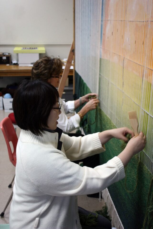 Group of women weaving threads affixed to a wall.